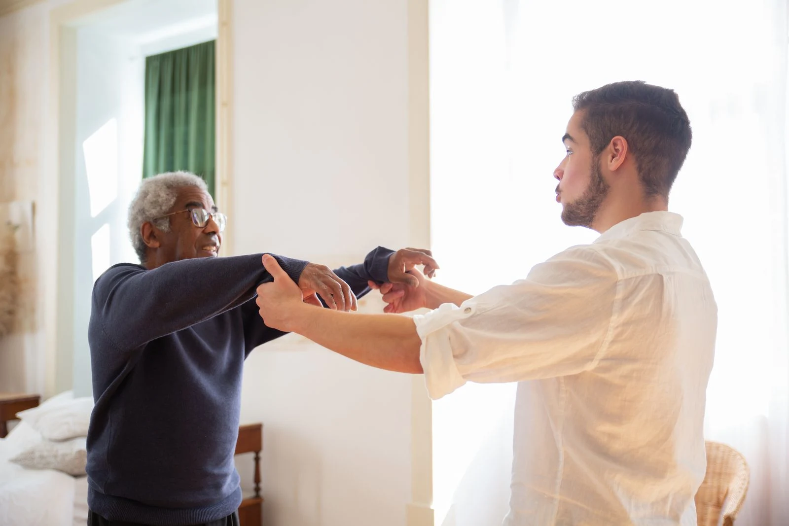A caregiver gently supporting an older man's arms in a bright, sunlit room at home.