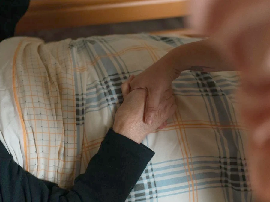 A caregiver's hand gently resting on an older person's hand on a soft blanket.